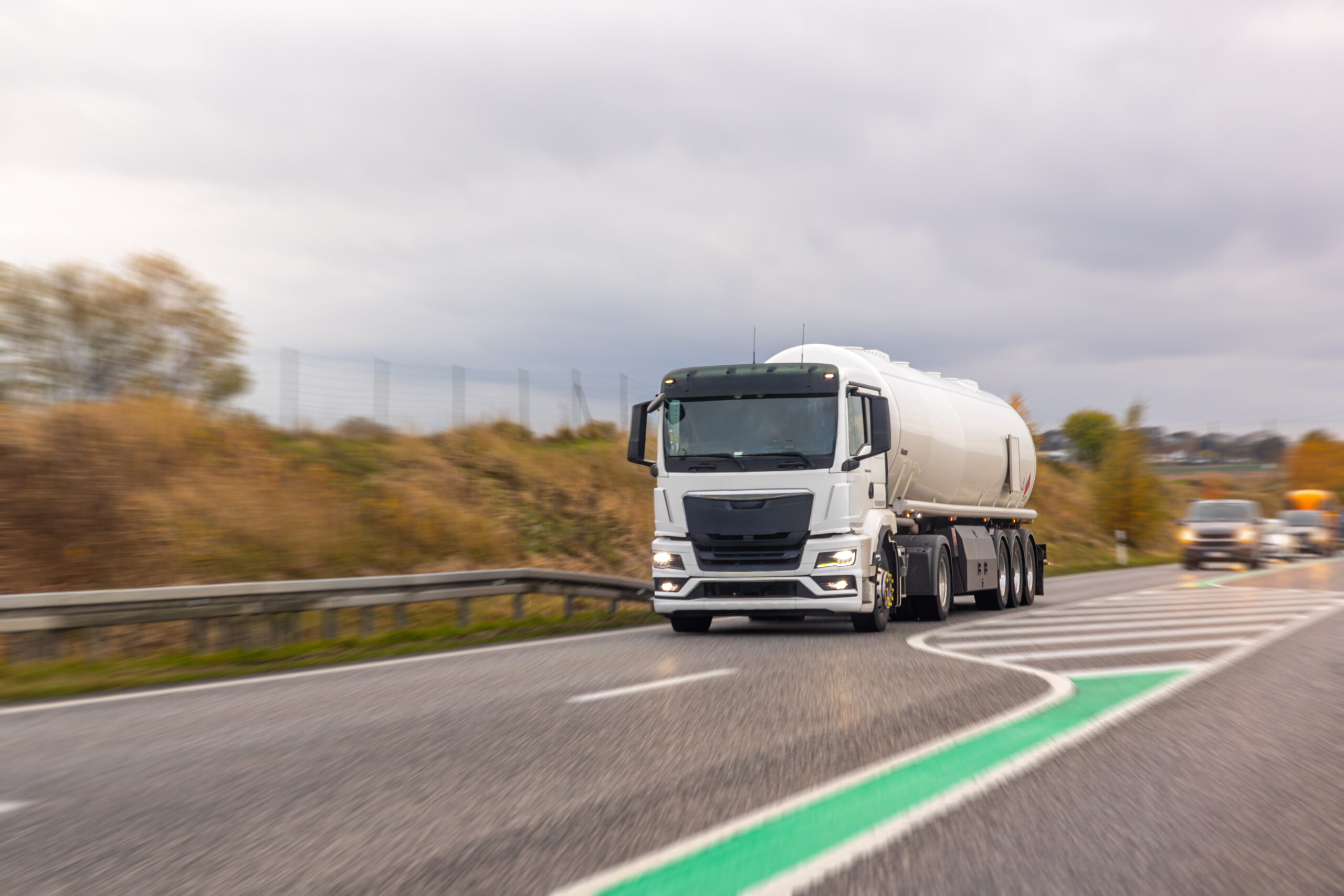 A white tanker truck takes a highway curve with motion blur, capturing speed and modern supply flow. Soft autumn colors and overcast sky add a cinematic transport mood.